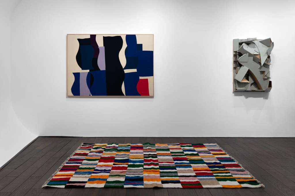 Gallery interior with abstract artworks on white walls and colorful striped rug on dark floor.