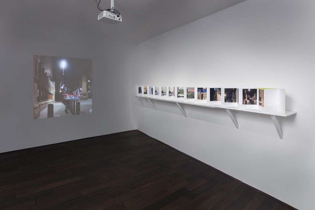 Gallery space with white walls, dark wood floor, projected image on left wall, and white shelf displaying framed photographs.