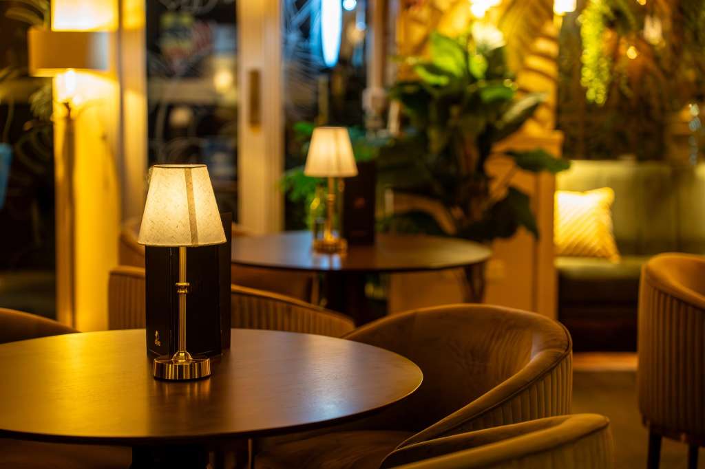 Intimate lounge area with round tables, table lamps, upholstered chairs, and tropical plants under warm lighting.