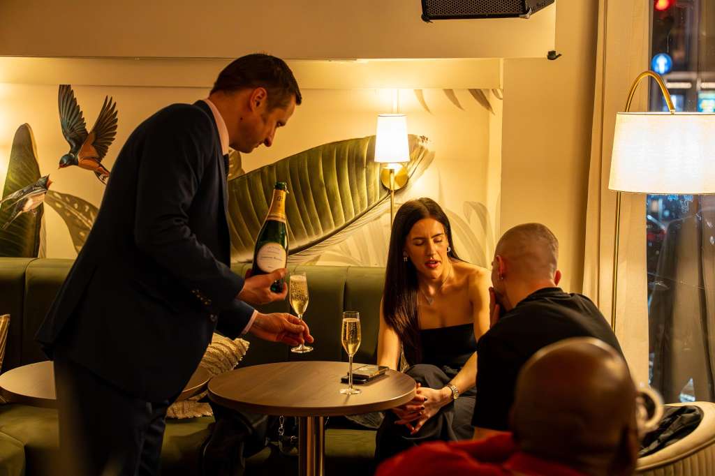 Lounge bar with guests seated on green banquette, server pouring champagne, warm lighting and botanical wall art.