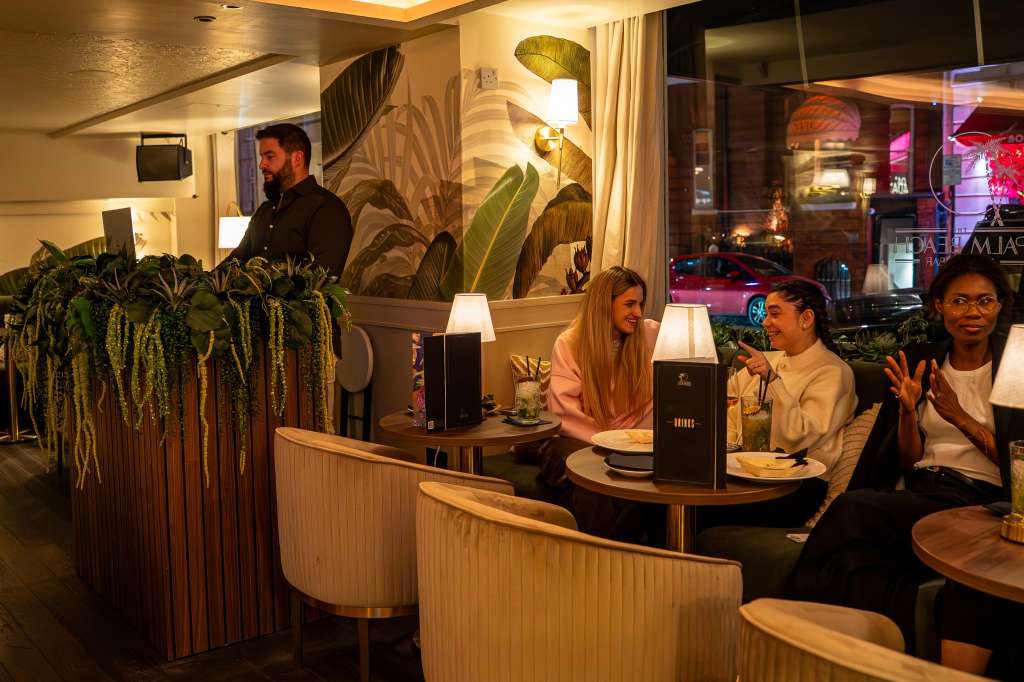 Lounge bar with curved seating, small tables, ambient lighting, tropical plant decor, and guests seated at tables.
