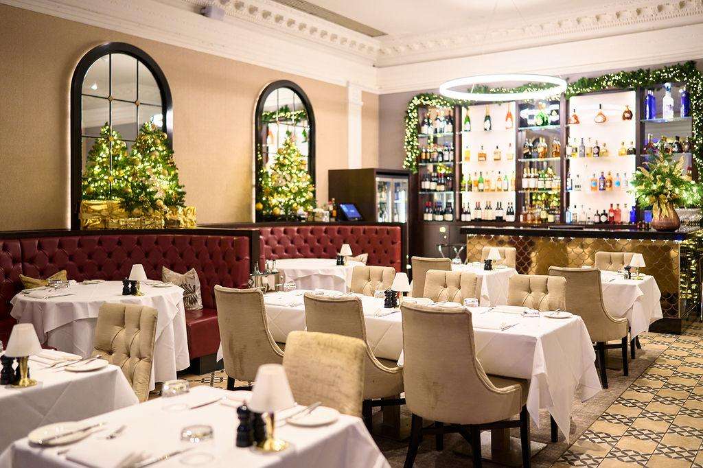 Fine dining room with round tables, white linens, cream chairs, burgundy banquette seating, and illuminated bar with bottles.