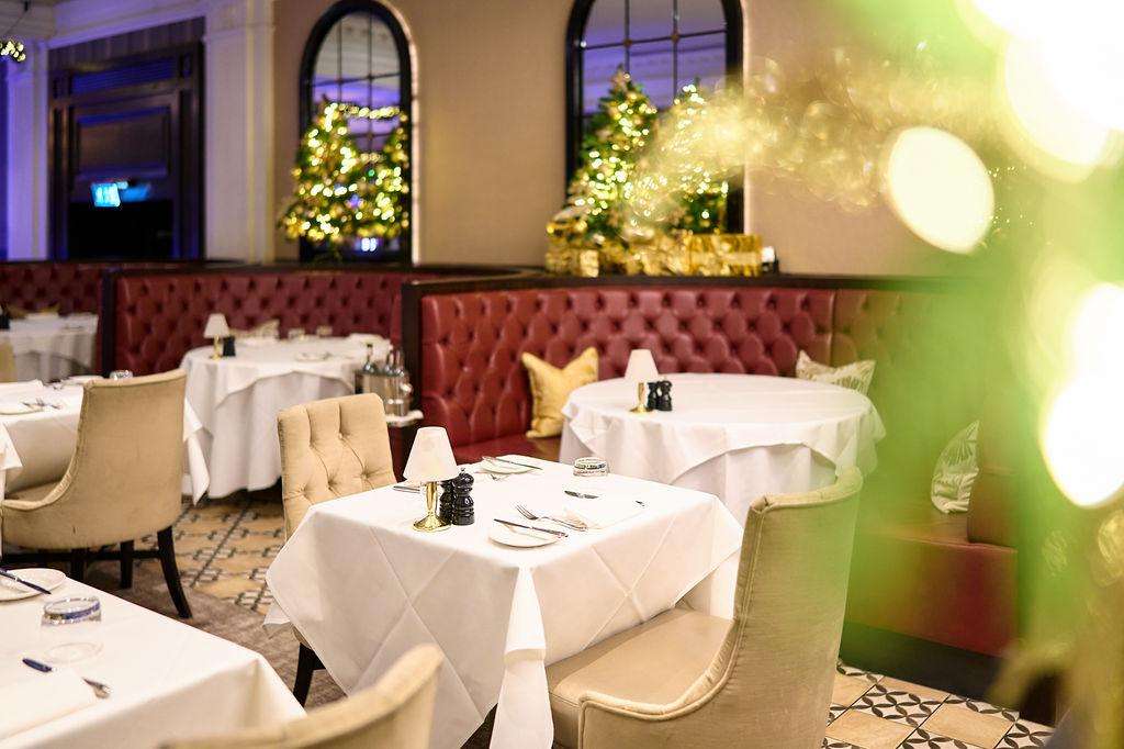 Dining room set for formal dinner with round tables, white linens, upholstered banquettes, and Christmas trees with warm lighting.