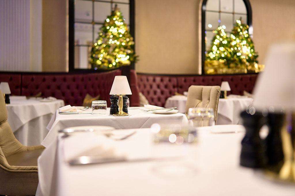 Dining room set for formal dinner with white tablecloths, place settings, and illuminated Christmas trees in background.