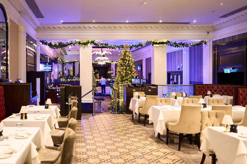Elegant dining room with round tables, white linens, cream chairs, and festive garland and gold tree decorations.
