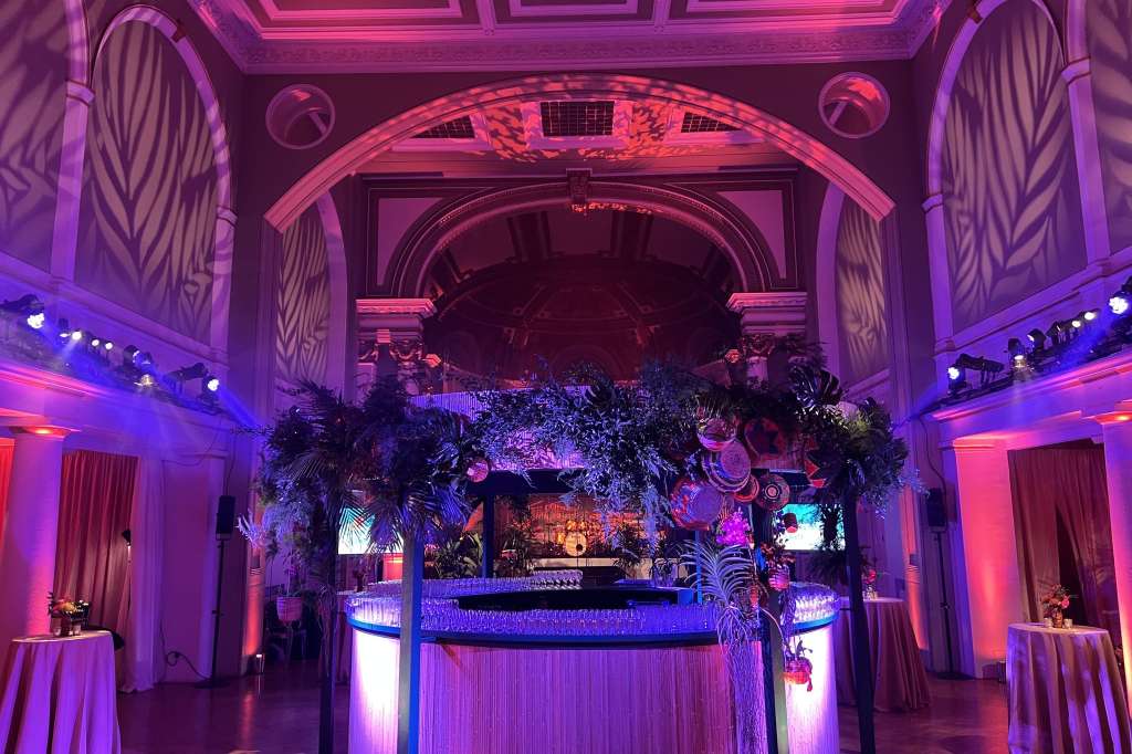 Soane Hall set up for an evening event with illuminated bar, tropical foliage installation, and dramatic purple and pink uplighting.