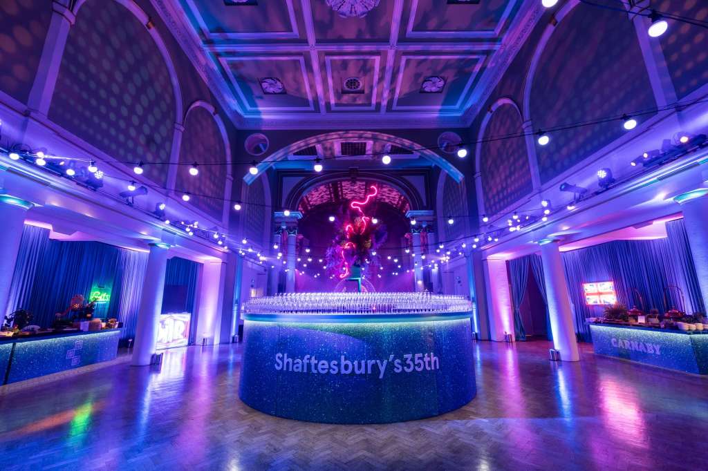 Grand hall with ornate ceiling, purple and blue ambient lighting, central bar, and neon signage for event branding.