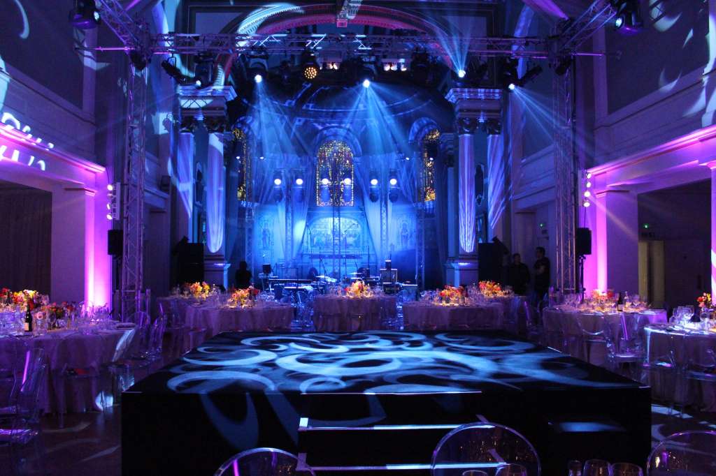 Soane Hall set up for an evening event with round tables, dramatic blue and pink uplighting, and a stage with performance setup.