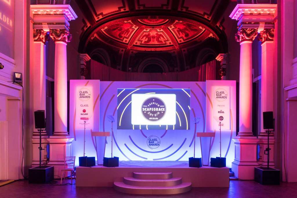 Grand hall with ornate dome, classical columns, and stage set with large screens and pink-purple lighting for an awards event.