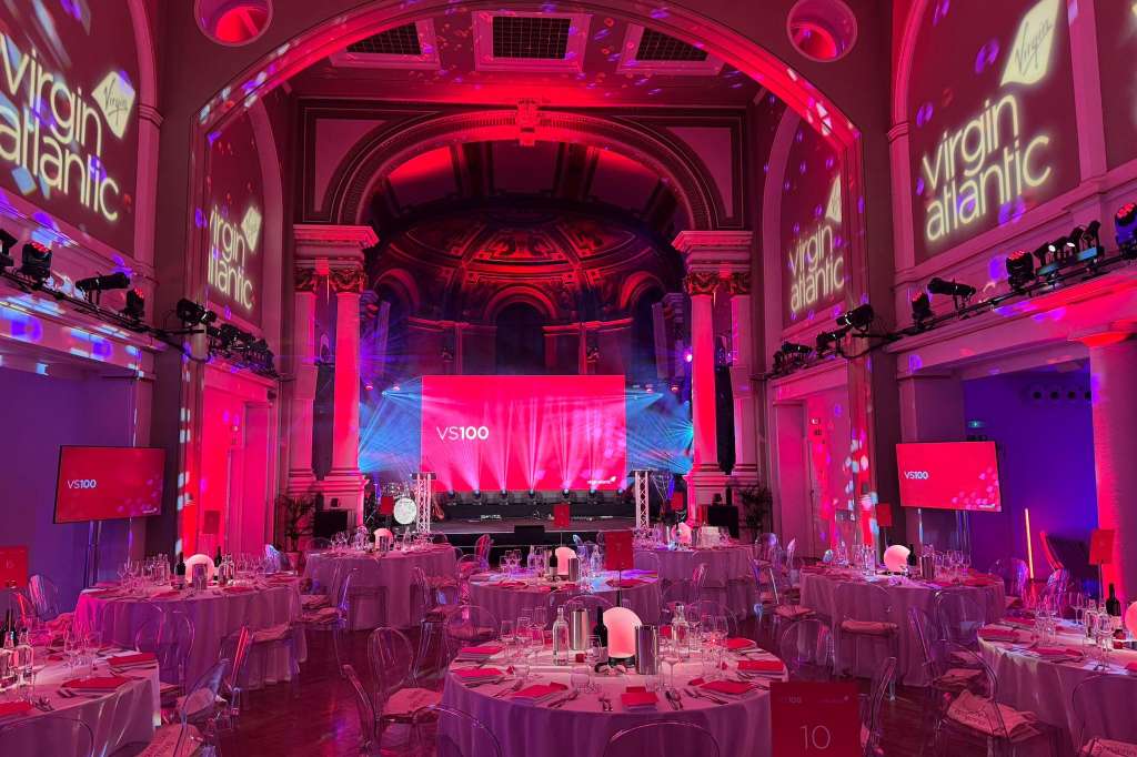 Grand hall with round tables, stage, and red-pink lighting. Virgin Atlantic branding visible on screens and signage.