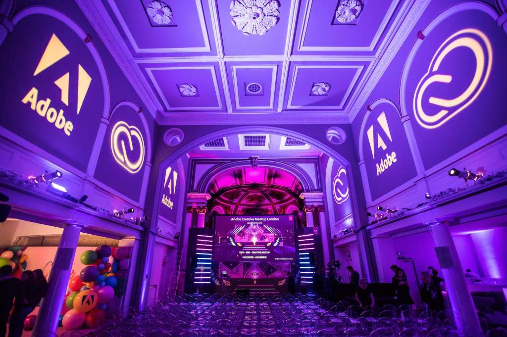 Gallery space with purple neon lighting, Adobe branding, geometric stage design, and attendees gathered for an event.