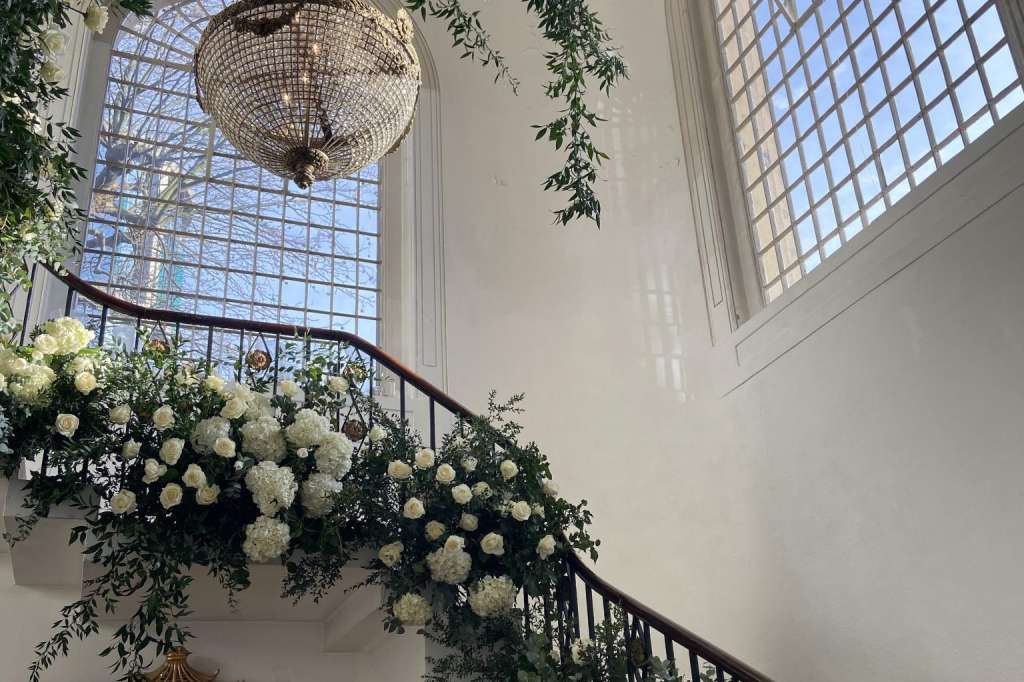 Soane Hall staircase decorated with cascading greenery, white hydrangeas, and a crystal chandelier under skylight.