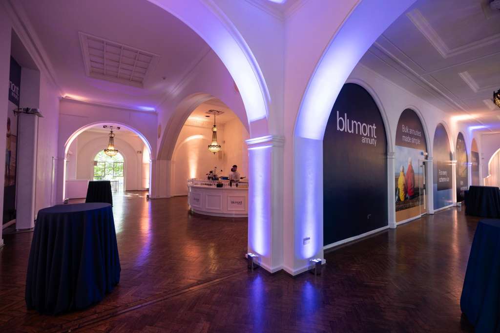 Gallery space with arched columns, atmospheric purple and blue lighting, high-top tables, and exhibition signage.
