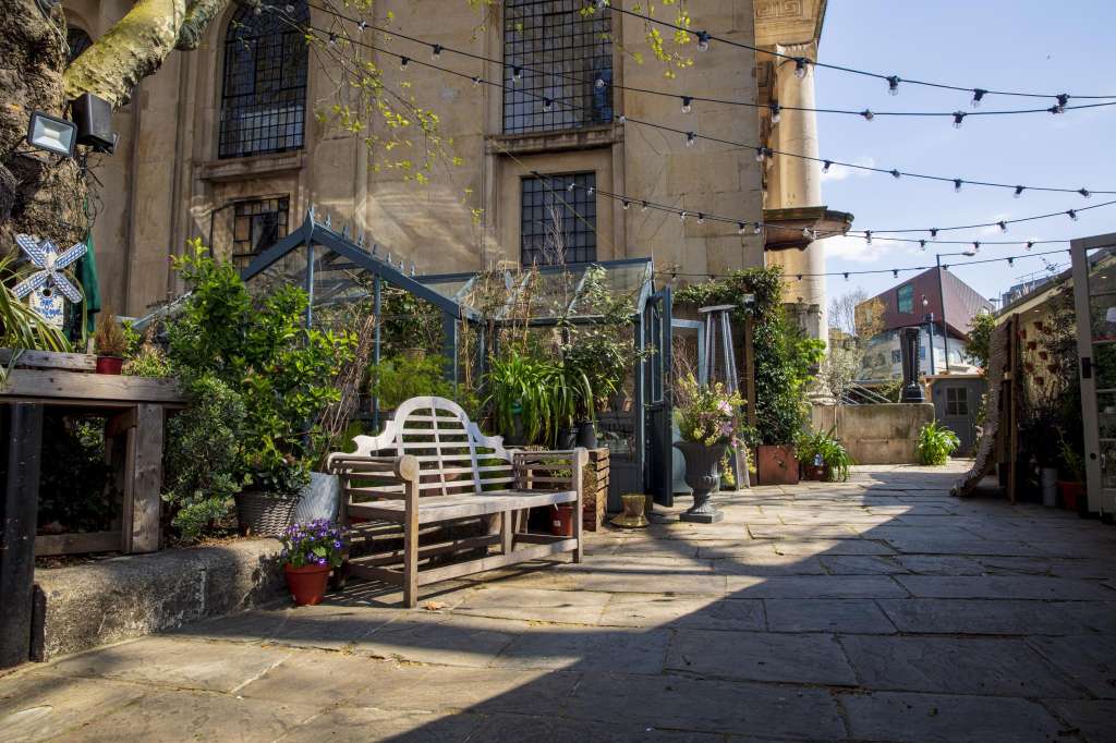 Outdoor courtyard with stone paving, planted containers, string lights, and wooden seating beneath historic stone building.