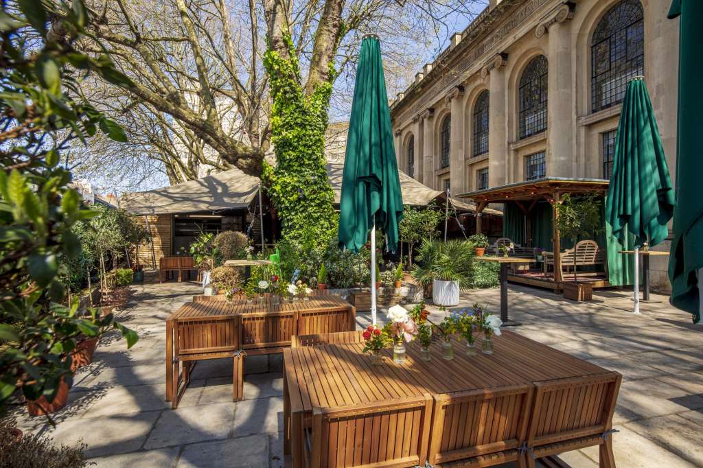 Outdoor courtyard with wooden seating, green umbrellas, ivy-covered columns, and flowering planters against a historic stone building.