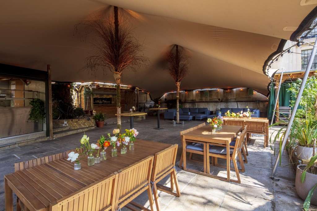 Rooftop terrace with wooden tables, blue seating, potted flowers, and decorative pampas grass under a canopy.