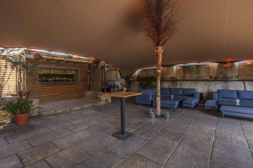 Rooftop terrace with bar, blue seating clusters, decorative tree, and ambient lighting at dusk.