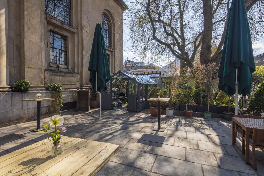 Outdoor courtyard with stone paving, wooden tables, closed umbrellas, potted plants, and historic building facade.