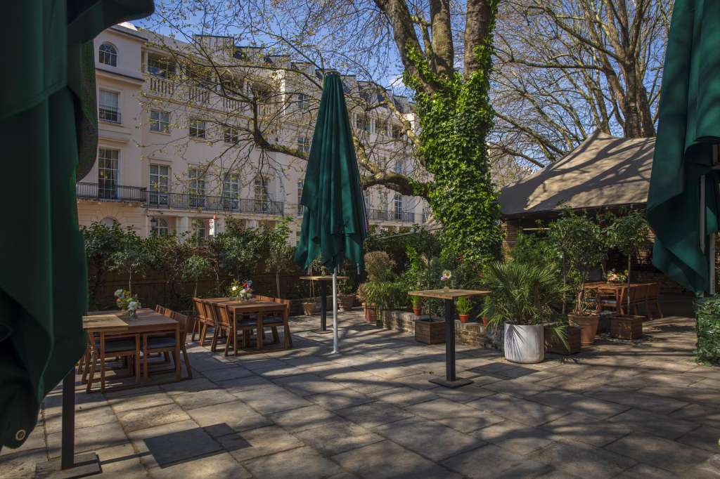 Outdoor courtyard with wooden dining tables, green umbrellas, ivy-covered trees, and Parisian architecture backdrop.