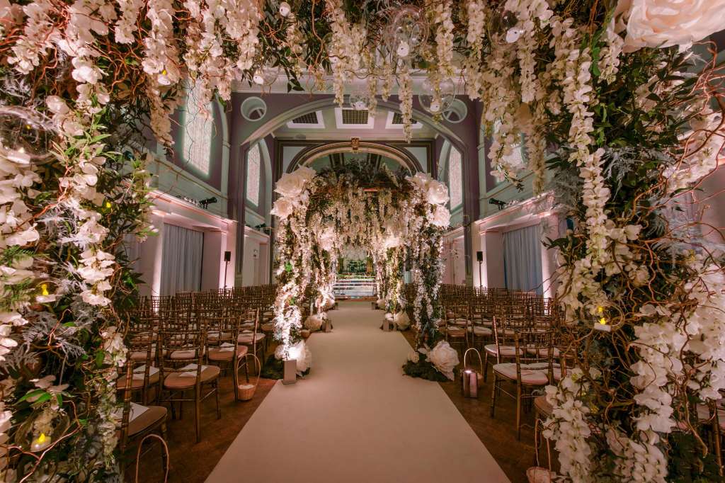 Soane Hall set up for a ceremony with elaborate floral installations, hanging white flowers, and a decorated aisle lined with gold chairs.