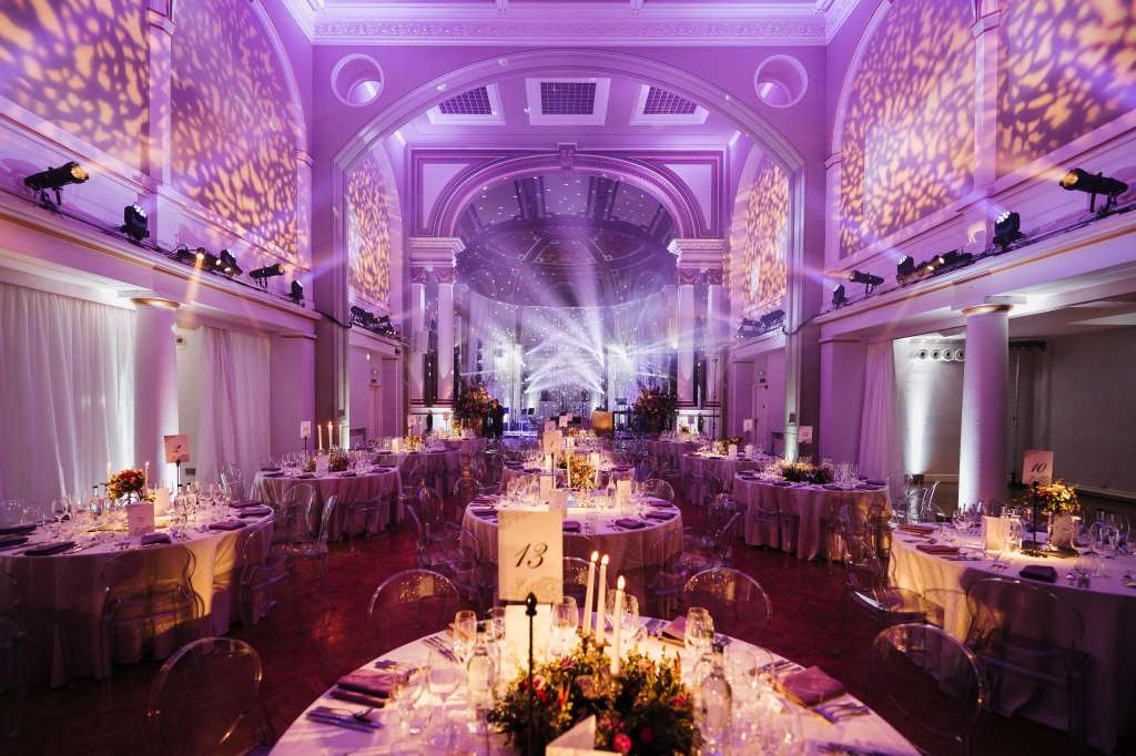 Soane Hall set up for a seated dinner with round tables, purple uplighting, ornate ceiling, and floral centerpieces.