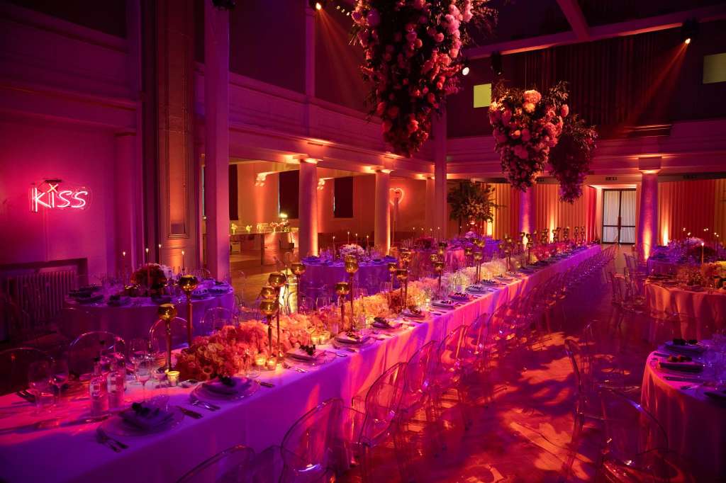 Soane Hall set for a seated dinner with long tables, dramatic pink and purple uplighting, hanging floral installations, and neon signage.