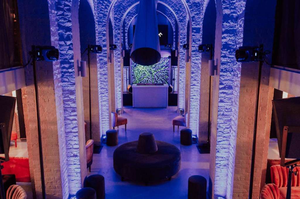 The Soane Hall with dramatic blue uplighting on vaulted brick arches, black seating pods, and red accent chairs arranged for an event.