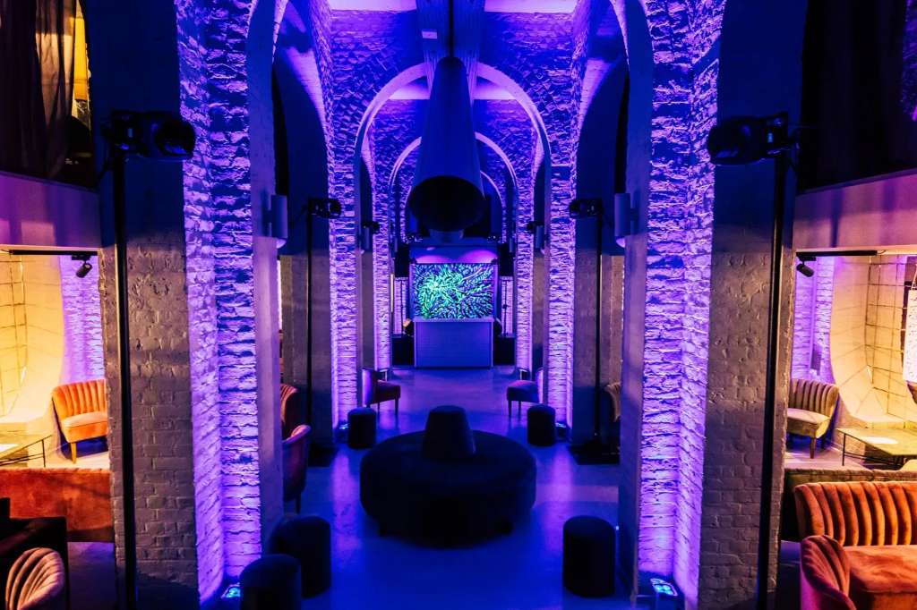 Gallery space with dramatic purple uplighting on exposed brick columns, modern seating, and digital display screens.