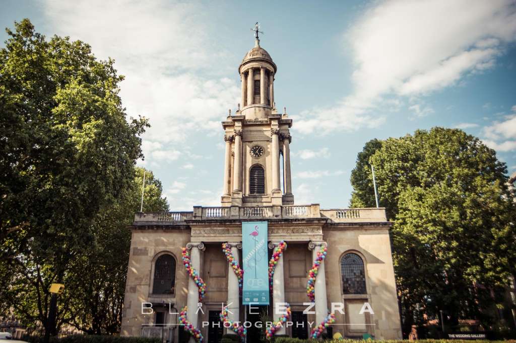 Exterior of historic stone building with clock tower, flanked by green trees and decorated with colorful floral garlands.