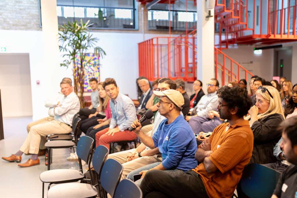Cormack room with seated audience facing a presenter, modern industrial space with red staircase and plant decor.
