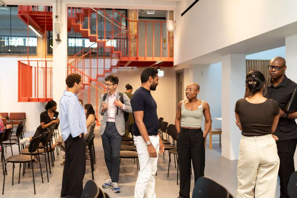 Modern industrial space with red staircase, groups of people standing and conversing, black chairs arranged around perimeter.