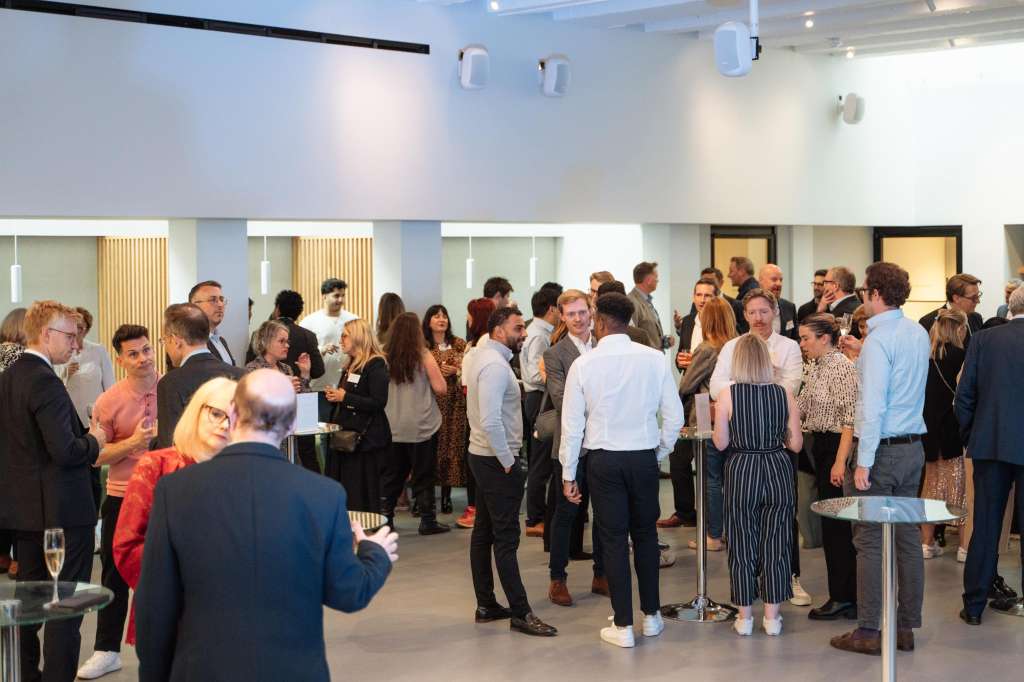 Modern event space with standing guests mingling, high ceilings, pendant lighting, and glass partitions visible in background.