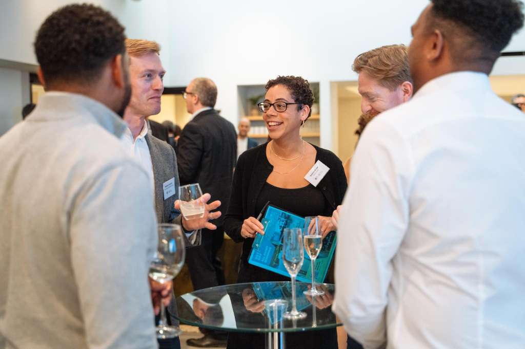 Group of professionals mingling at a standing reception with drinks and name badges in a modern interior space.