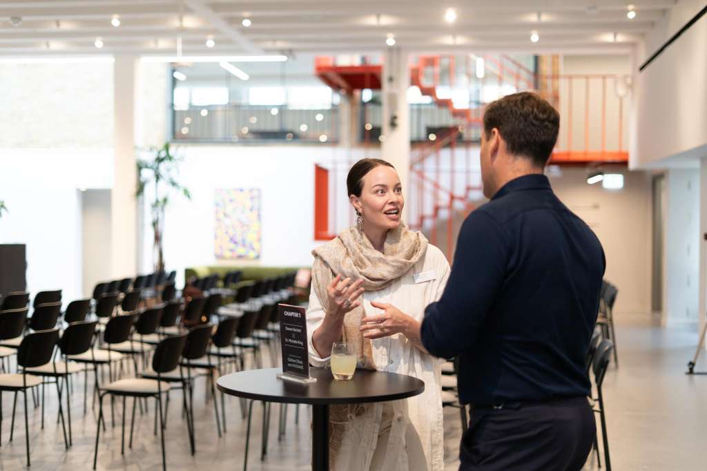 Modern meeting space with rows of black chairs, two people conversing at a high table, bright lighting, and contemporary artwork on walls.
