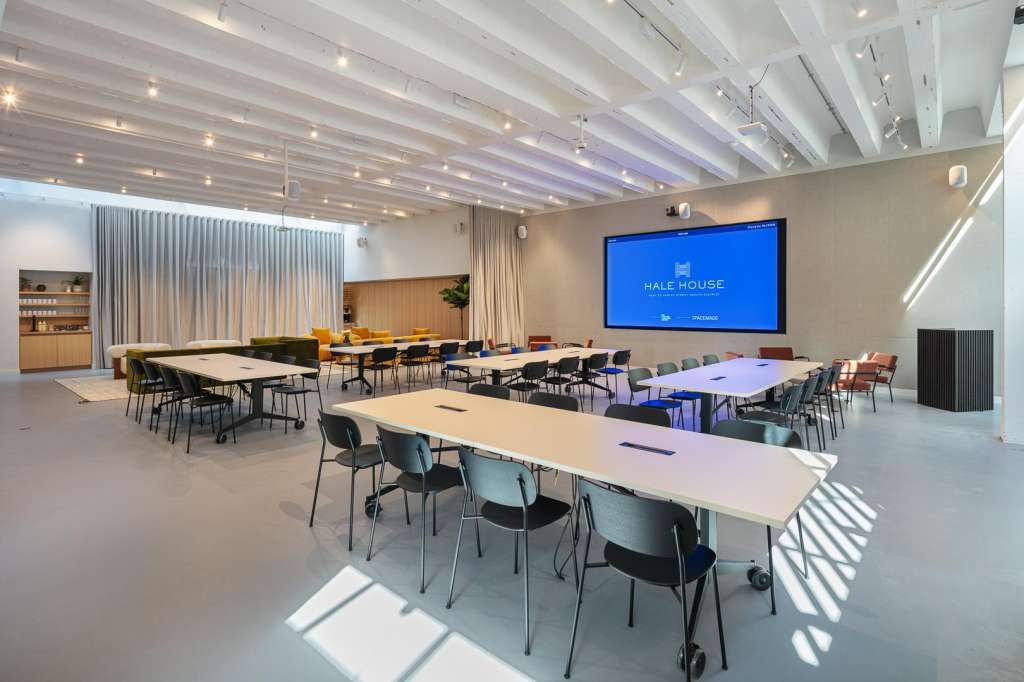 Cormack room set up with multiple rectangular tables, chairs, and a large blue screen displaying Hale House branding.