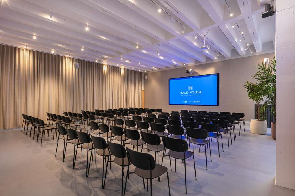 Cormack room set up with rows of black chairs facing a large blue screen displaying Hale House branding, white ceiling beams, and warm curtains.