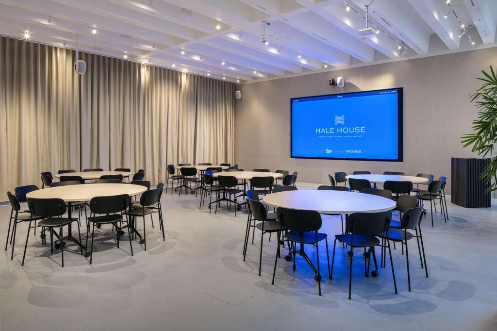 Modern meeting room with round tables, black chairs, projector screen displaying Hale House branding, and ambient blue lighting.