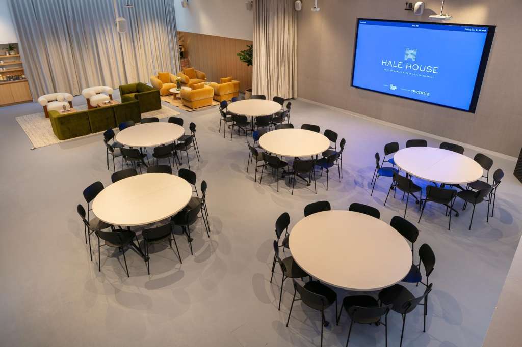 Cormack room set up with multiple round tables, black chairs, lounge seating, and large display screen showing Hale House branding.