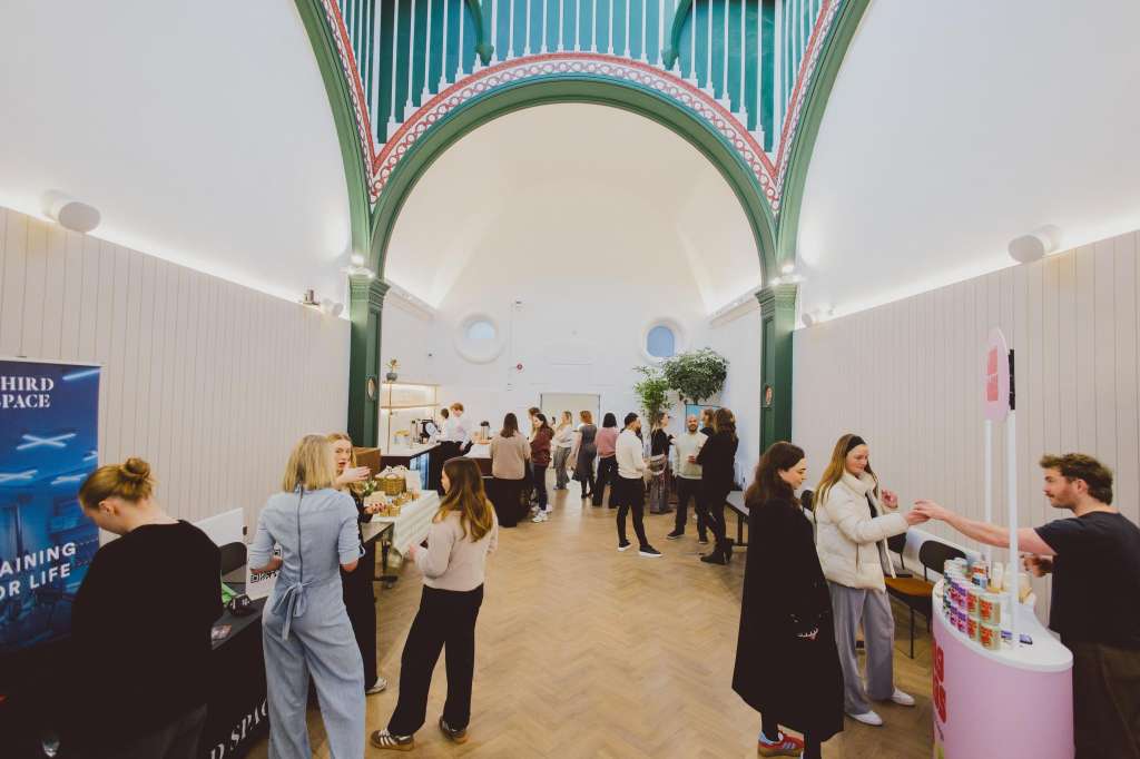 The Chapel interior with vaulted ceiling, green pillars, and decorative arch. Multiple guests mingling around exhibition stands and displays.