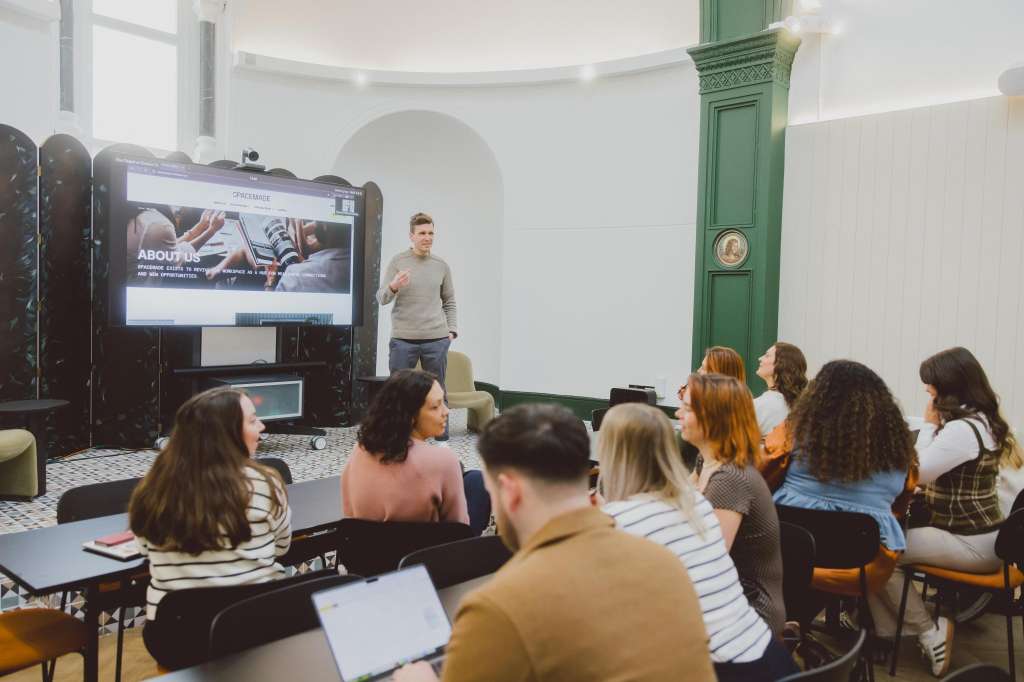 The Chapel set up for a presentation with attendees seated facing a large screen and standing presenter.