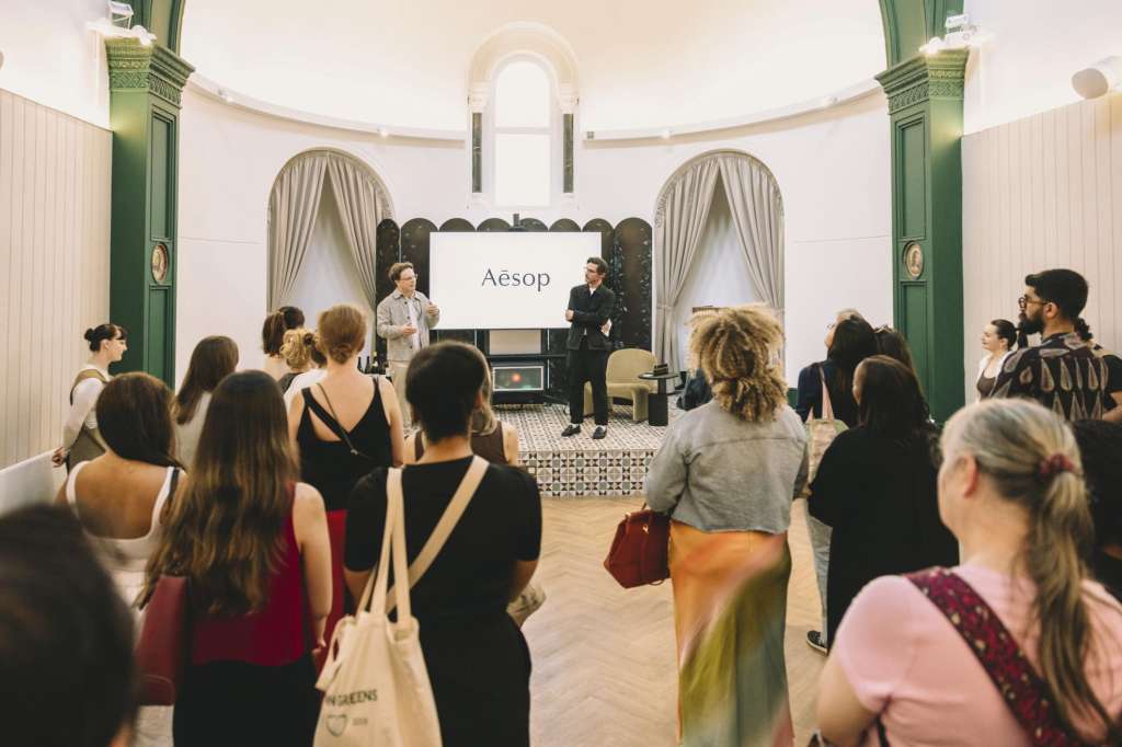 The Chapel set up for a presentation with audience seated facing a stage displaying an Aesop banner and two speakers.
