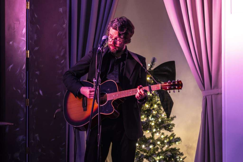 Musician performing with acoustic guitar on stage in The Chapel, lit by purple ambient lighting, with illuminated Christmas tree visible.