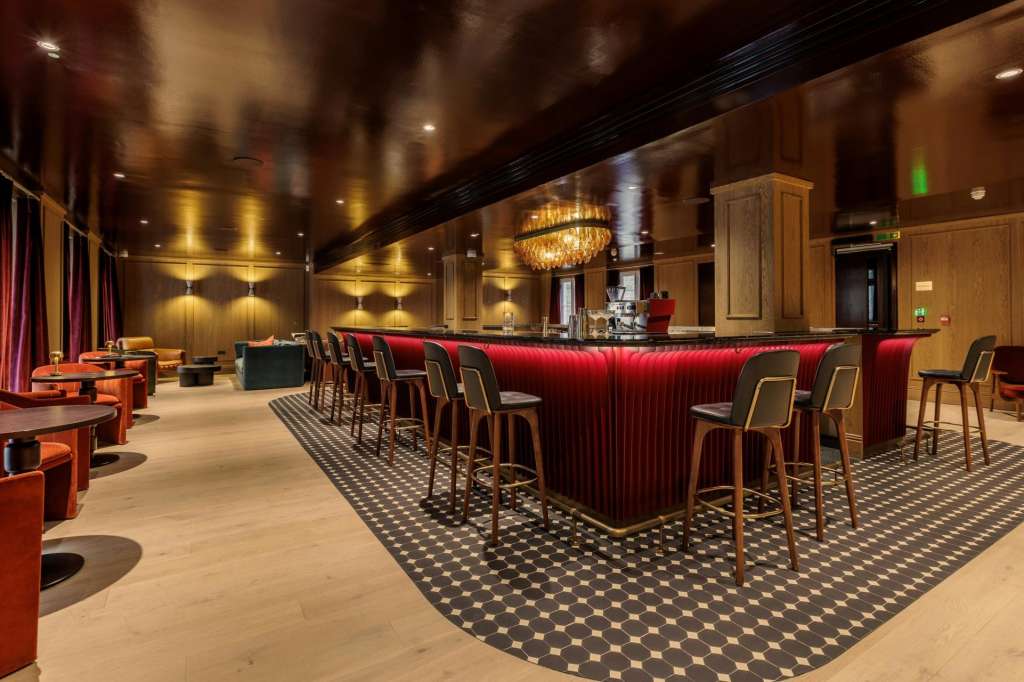 Bar area with red upholstered counter, wooden stools, warm lighting, and geometric floor pattern.