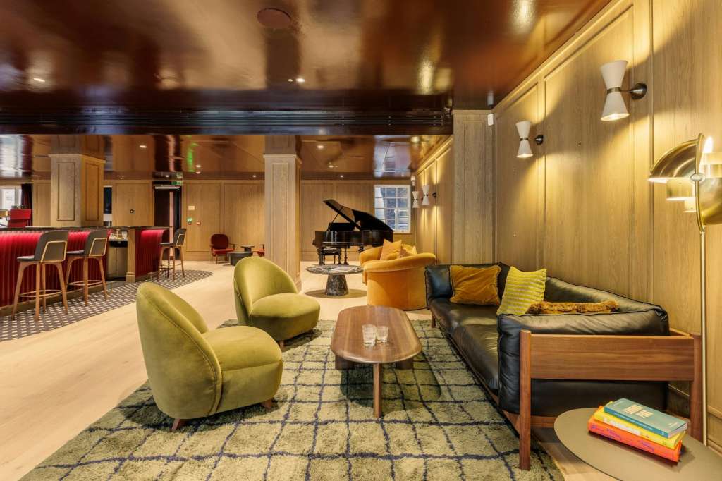 Modern lounge with yellow armchairs, black leather sofa, grand piano, bar area, and warm wood paneling throughout.