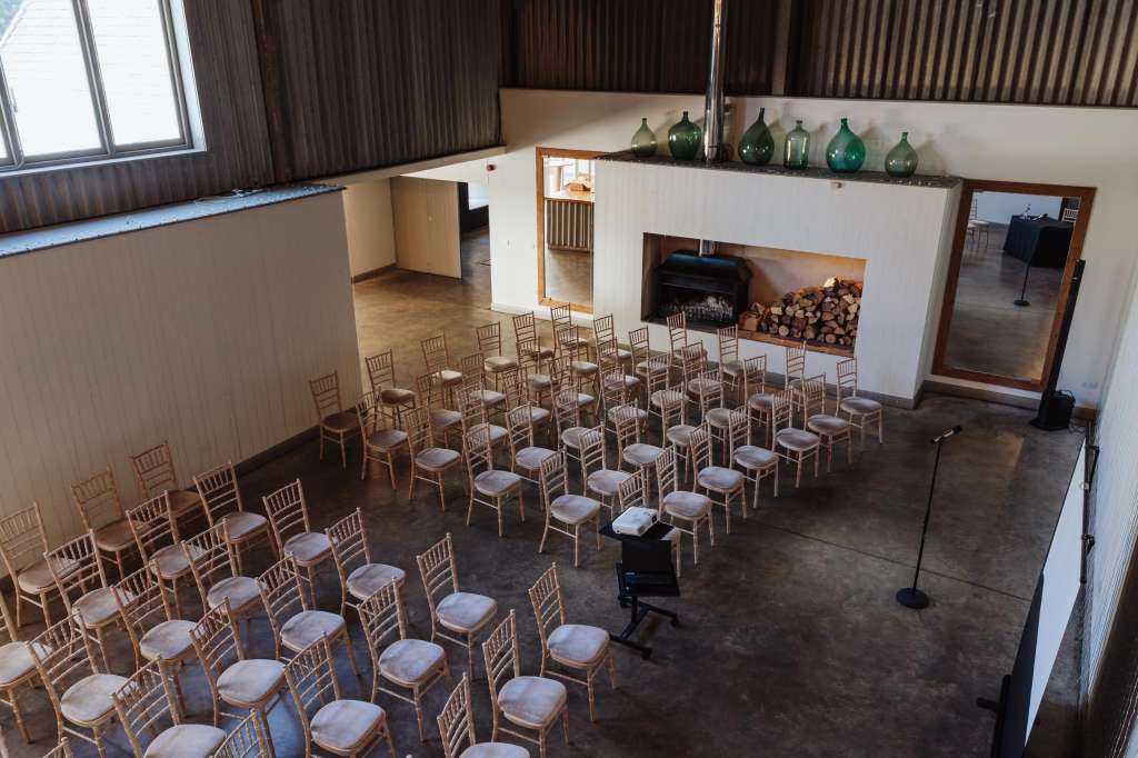 Industrial space with rows of wooden chairs with cushions arranged in theater-style seating, fireplace, and decorative bottles.