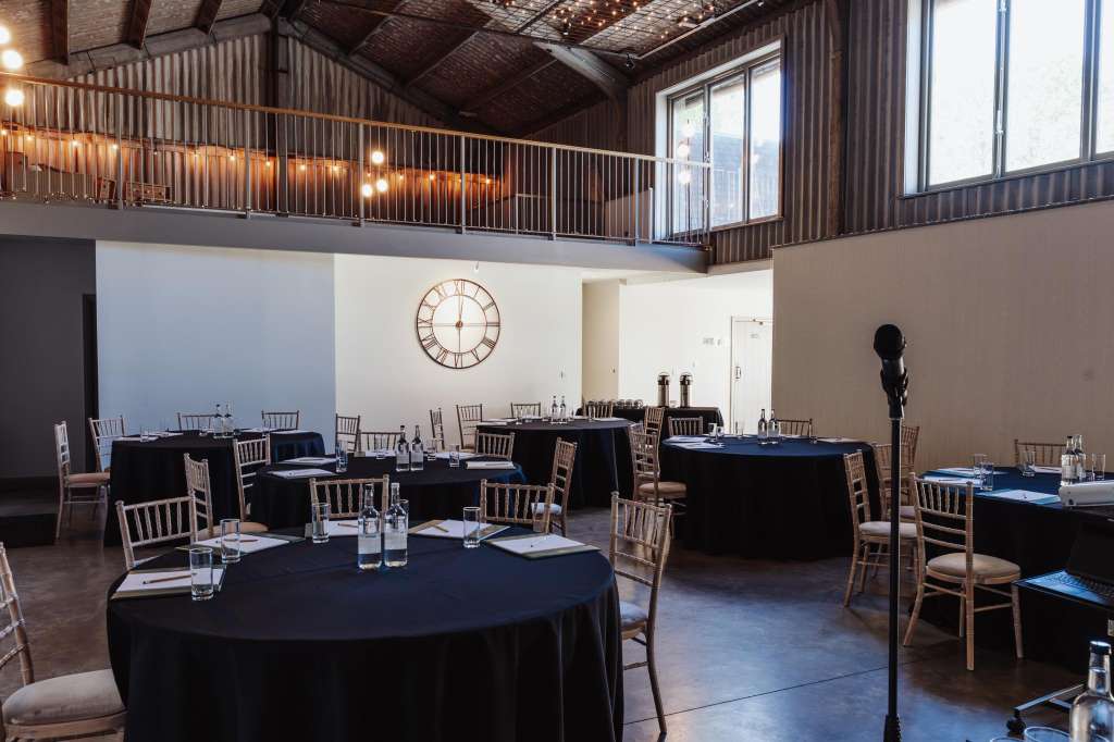 Industrial venue with round tables dressed in black linens, wooden chairs, and place settings. Mezzanine with string lights above.
