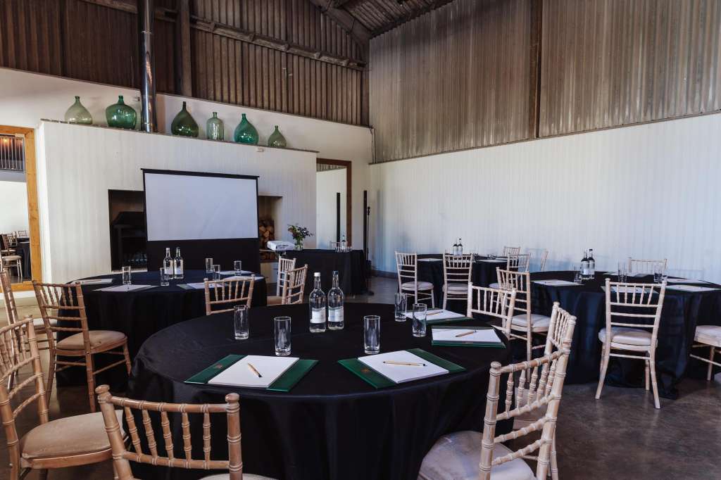 Industrial barn space with round tables dressed in black linens, chiavari chairs, projection screen, and water bottles set for a conference or corporate event.