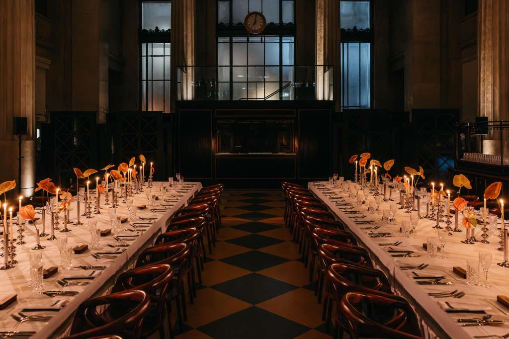Main Hall set for a formal seated dinner with long tables, place settings, orange floral centerpieces, and candlelight.