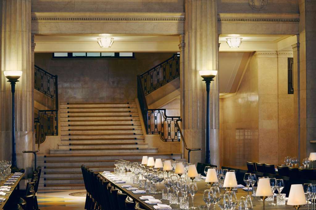 Main Hall set for a formal seated event with long tables, place settings, glassware, and table lamps; grand staircase visible in background.