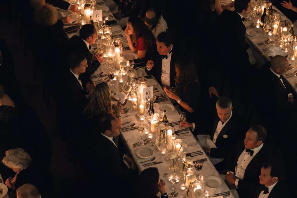 Long banquet table with candlelit place settings and formally dressed guests standing and mingling in dim lighting.